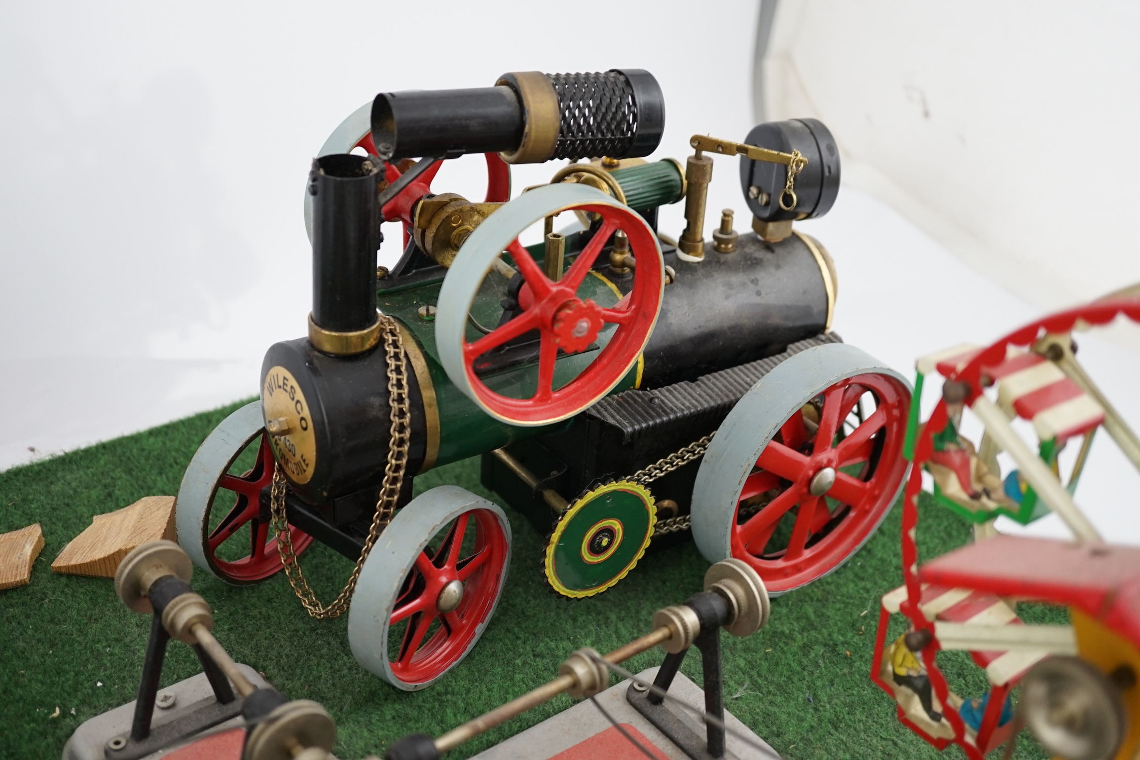 A Wilesco live steam fairground diorama, comprising a D430 pellet fired traction engine, powering a pulley and gear assembly, in turn running three tinplate fairground rides, mounted on a plywood sheet topped with grass